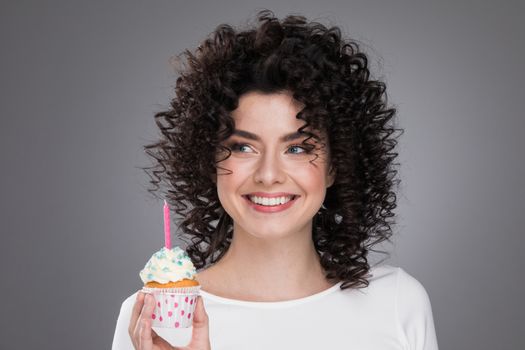 Cheerful beautiful curly young woman making a wish holding birthday cupcake with candle