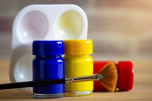 Poster colour bottle, paintbrush and palette on wooden table with brick wall background and morning sunlight.