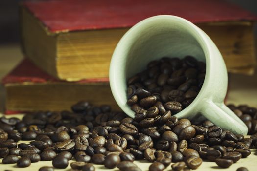 Coffee bean in tilted ceramic cup and old book laid behind on wooden table with morning light.