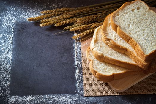 Bread on a wooden cutting board and the wheat grains placed beside With copy space frame of wheat flour scattered. Breakfast on the marble table and morning sunlight.