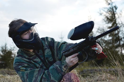 Paintball sport player girl in protective camouflage uniform and mask with marker gun outdoors