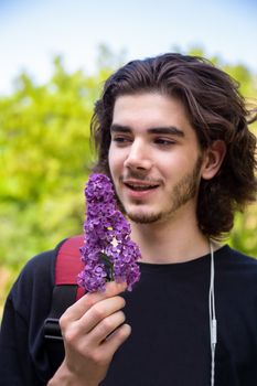Young brunette guy smelling fresh flowers on lilac branch and looking at camera on blurred background