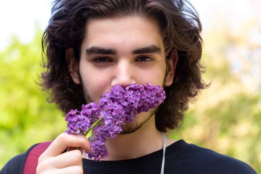 Young brunette guy smelling fresh flowers on lilac branch and looking at camera on blurred background