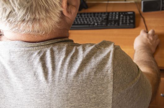 An elderly gray-haired man uses a computer mouse, work at home for the disabled, training pensioners to work on a PC, a view from the back.