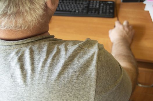 An elderly gray-haired man uses a computer mouse, work at home for the disabled, training pensioners to work on a PC, a view from the back.