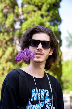 Young brunette man with long curly hair and sunglasses holds fresh flowers in his mouth on a lilac branch and looks away on a blurred background.
