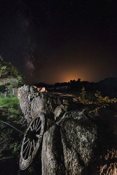 A wagon wheel is supported on the long wall of large stones of a farm. The dark night sky is full of stars and behind the mountains you can see the reflection of the lights of a nearby town