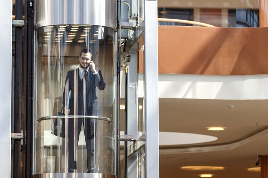Businessman in modern glass elevator talking by the phone and holding briefcase