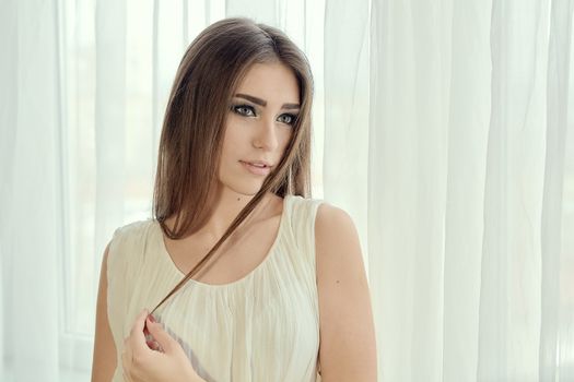 Cheerful smiling young white skin female with long brunette hair posing against the window in the morning, meeting a new day, dressed in a light transparent white tunic. Morning makeup.