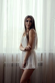 Cheerful smiling young white skin female with long brunette hair posing against the window in the morning, meeting a new day, dressed in a light transparent white tunic. Morning makeup.