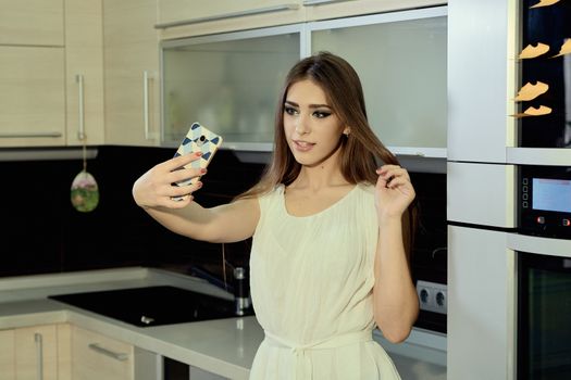 Cheerful smiling young white skin female with long brunette hair posing on the kitchen, makes selfie on the smartphone, dressed in a light transparent white tunic. Morning makeup.