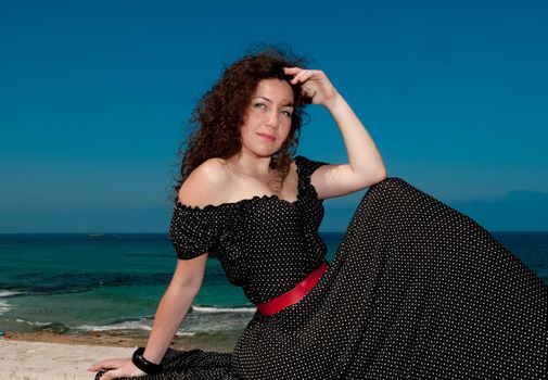 Young, beautiful woman in a black  dress with white polka dots posing against the sea