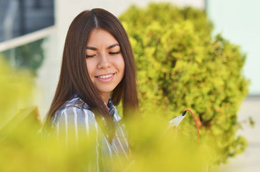 ? young girl of Asian appearance listening to music on the street, sitting on a bench near the green bushes