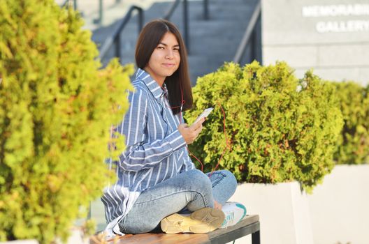 Young beautiful is asian girl sitts on the bench near green tress and listen to music. Spring sunny day