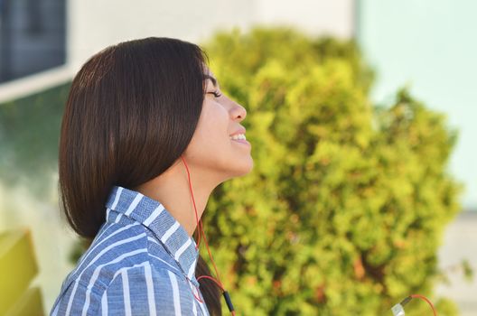 Young beautiful is asian girl sitts on the bench near green tress and listen to music. Spring sunny day