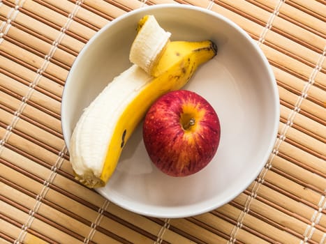 Banana and apple inside white bowl, ready for light meal, on wooden table.