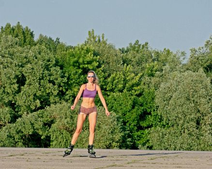 Blonde girl in sunglasses on roller-skates in sunny weather on an asphalt path on the green trees background