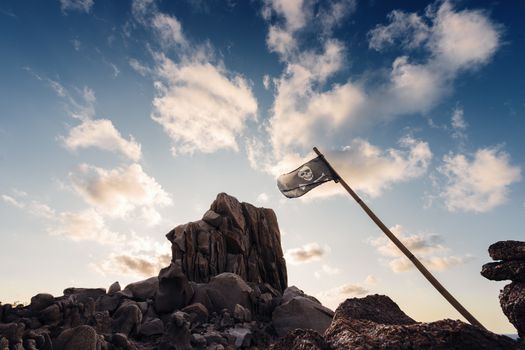 jolly roger flag flies pinned between the rocks of the pirate island in front of a few clouds in the clear blue sky, Capo Testa, Santa Teresa di Gallura, Olbia Tempio, Sardinia, Italy