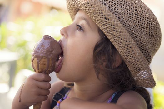 Little girl with a straw hat and a summer dress enjoys the summer heat eating a refreshing cone of chocolate ice cream, it creamy melts in her hand while she licks it with her tongue