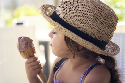 Little girl with a straw hat and a summer dress enjoys the summer heat eating a refreshing cone of chocolate ice cream, it melts in her hand while she looks at it with desire