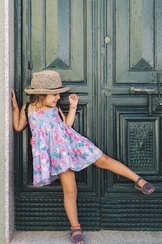 Little girl poses raising one leg in front of the green door of the house, she wears a lilac summer dress of flowers and a straw hat, vertical image