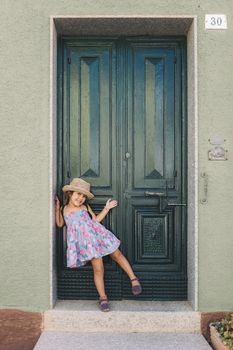 Little girl poses amused and smiling in front of the green door of her house, she wears a lilac summer dress of flowers and a straw hat, vertical image