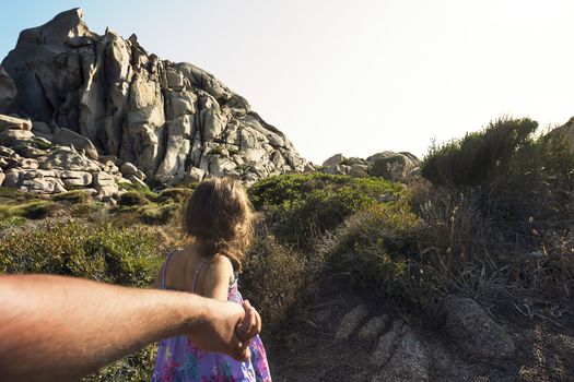 Kid strolling through the bushes holding her father's hand, in the background there are rocky mountains and the sky shines clear, subjective shot, Capo Testa, Santa Teresa di Gallura, Sardinia, Italy