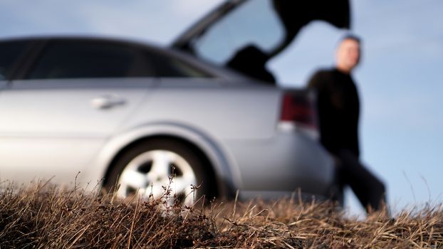 Sde view of young businessman in eyeglasses sitting on car trunk at nature - blurred background for banner