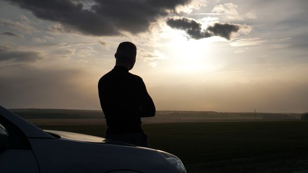 Silhouette of the young man standing near to car and looking at a sunset