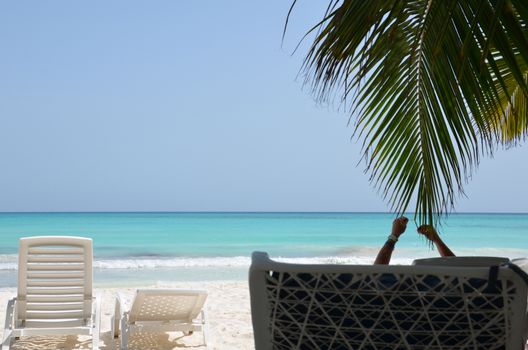 Beautiful sea and white sand. A man lies on the deck chair, holding up their hands, in the shade of palm trees on the beach