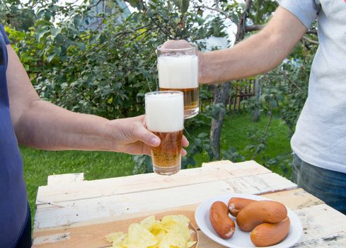 hands holding glasses with beer, chips and sausages lying on a light wooden boards, people relax in the weekends