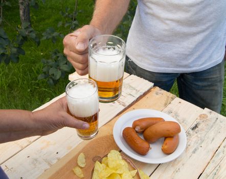 hands holding glasses with beer, chips and sausages lying on a light wooden boards, people relax in the weekends