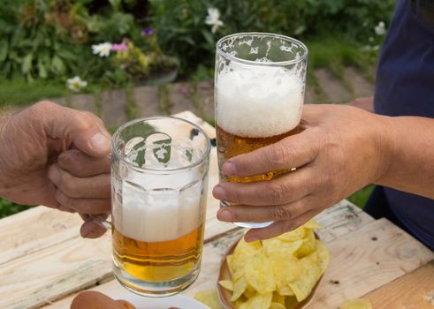 hands holding glasses with beer, chips and sausages lying on a light wooden boards, people relax in the weekends