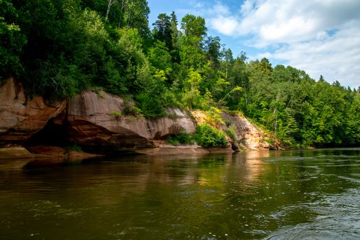 Landscape with cliff near the river Gauja, forest and clouds reflection in water. Cave in cliff. The Gauja is the longest river in Latvia, which is located only in the territory of Latvia. 

