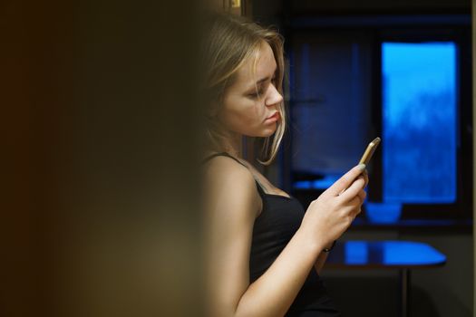 Indoor shot of European young woman holds modern phone, dressed in casual clothes, uses internet for networking and communication, stands near wall