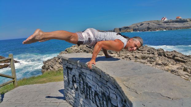 man in white shorts and white t-shirt does gymnastic exercise on a stone wall against a blue sea in Soto de Marina, Spain