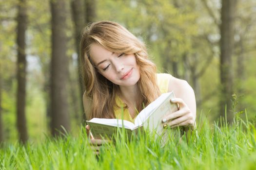 Pretty girl laying on the grass and reading a book in spring park and smiling