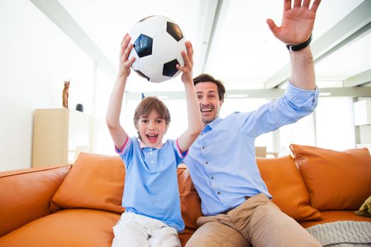 Portrait of happy boy watching soccer match with father on sofa at home