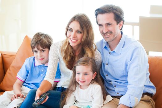 Beautiful young parents and their children are watching TV, eating popcorn and smiling while sitting on couch at home