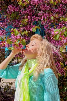 A beautiful young blond woman with curly hair stands near a flowering tree on a warm spring or summer day and sniffs purple flowers with eyes closed. Copy space for inscription.