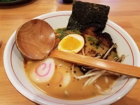 bowl of spicy asian noodles with egg, seaweed, and wooden spoon