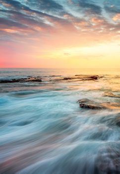 Incoming waves wash over rocks at sunrise which lights up the sky in hues of yellow orange and pinky reds