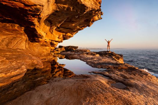 Female standing on the undercliff ledge enjoing the coastal views of Sydney in some early morning light which is highlighting the eroded rock details and reflecting the sky in a pool of water