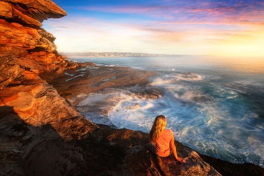 Female watches the ocean wash up and cascade like a waterfall and toss and swirl around ocean rocks with views to Bondi in a soft misty early morning light