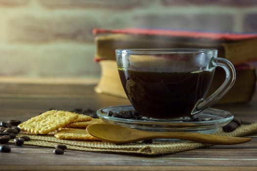 Black coffee in clear glass cup and coffee beans with crackers and old book on wooden table.