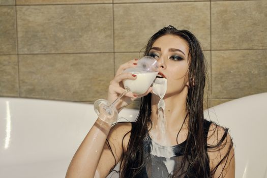 Young woman pours milk on herself. Woman with smeared makeup dressed in the black bathing suit posing in the bath of milk. Conceptual fashion photography for design. Young woman for lifestyle design.