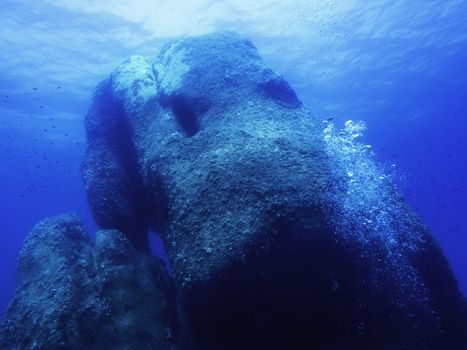 Underwater stone, Pietra di Saragossa, Sardinia. The large rock reaches from the seabed to the surface illuminated by the sun. Small fish swim around, some bubbles go up on one side