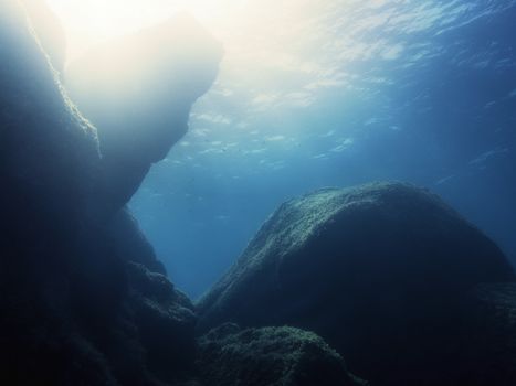 The dark rocks of the seabed are backlit by sunlight, this is reflected in the golden surface of the crystal blue water