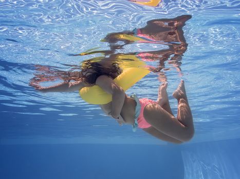 underwater view of a little girl swimming relaxed with his yellow float in the pool, summer sunlight reflected on the blue water surface