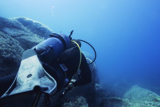 person diving near the rocks of the sea floor of a blue and turquoise sea, the stones are covered with small seaweed. The diver turns his back to the camera and one of his fins is in the foreground